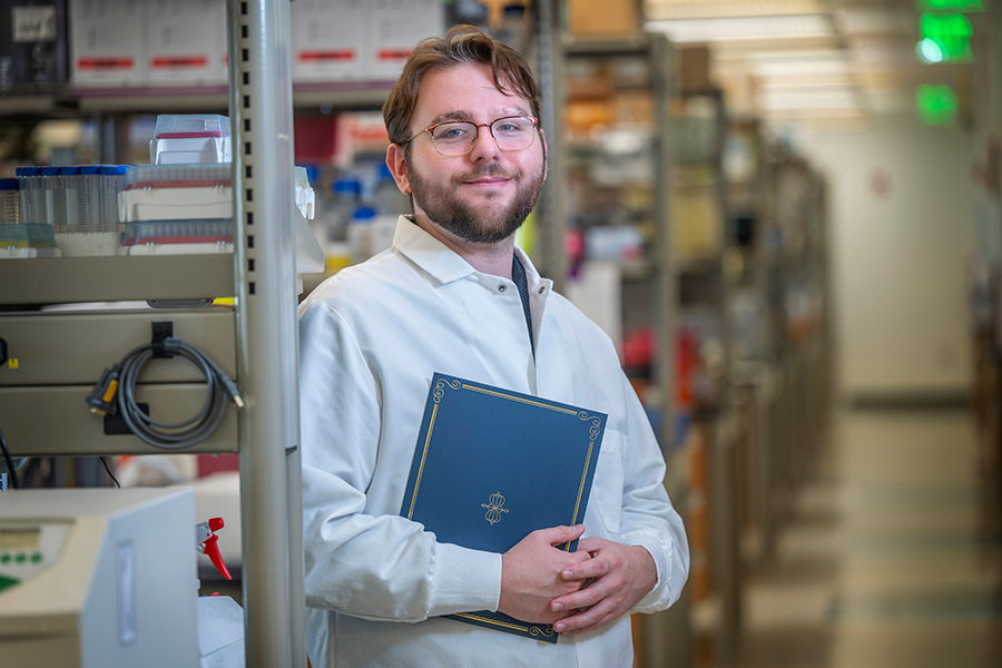 A male college student wearing a scientific lab coat and holding a folder stands in a large science lab.