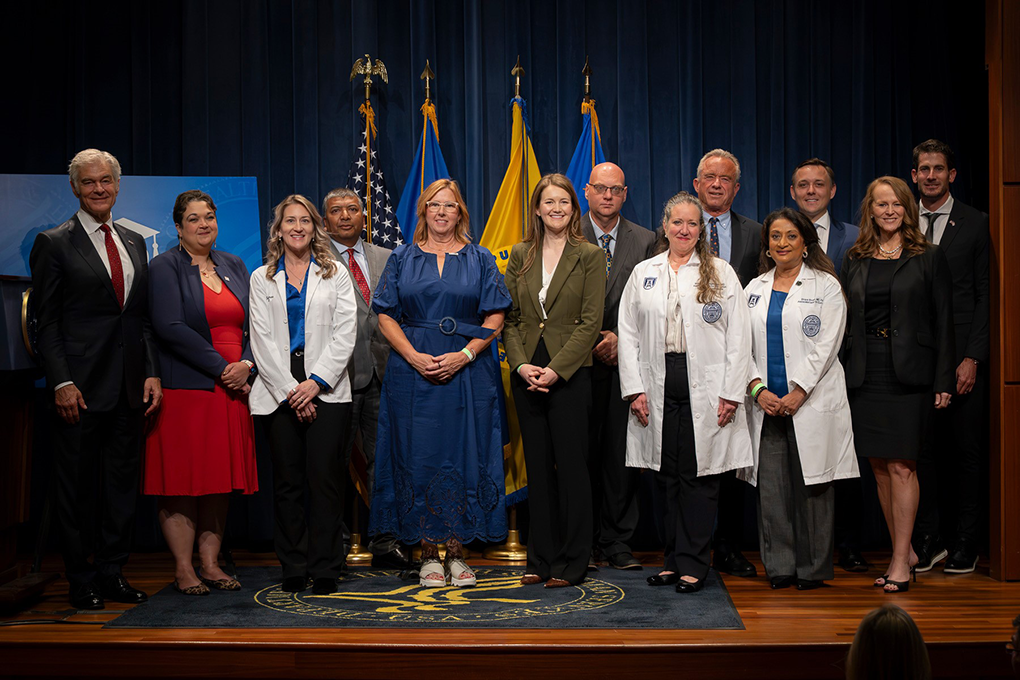 Men and women in business dress and white medical coats stand in front of flags.