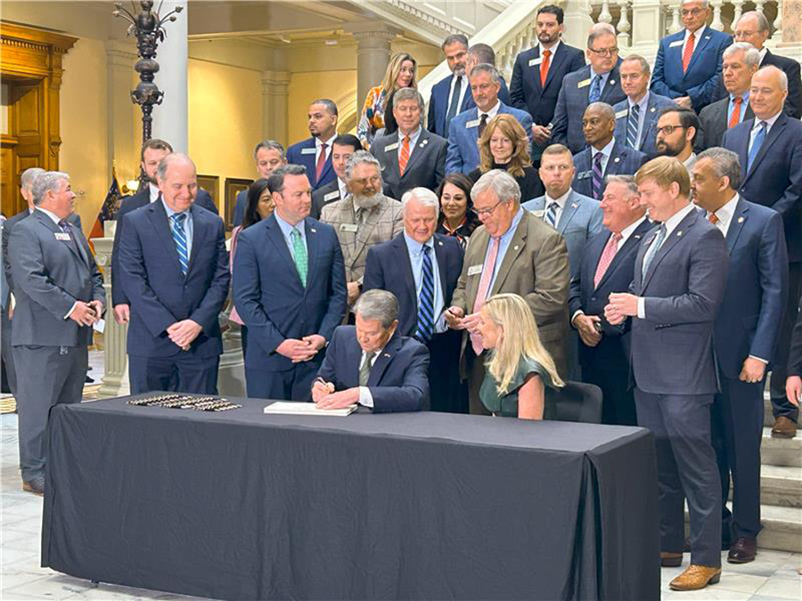 A large group of people gather around a man and a woman as the man signs a piece of paper. They are in a large atrium of a state capitol building.