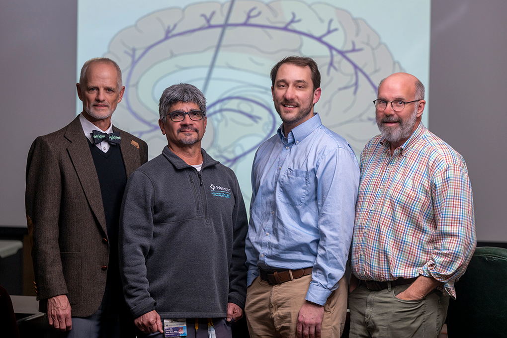 A group of four male neuroscientists stand in front of a backdrop with a picture of an animated brain on it and look at the camera.
