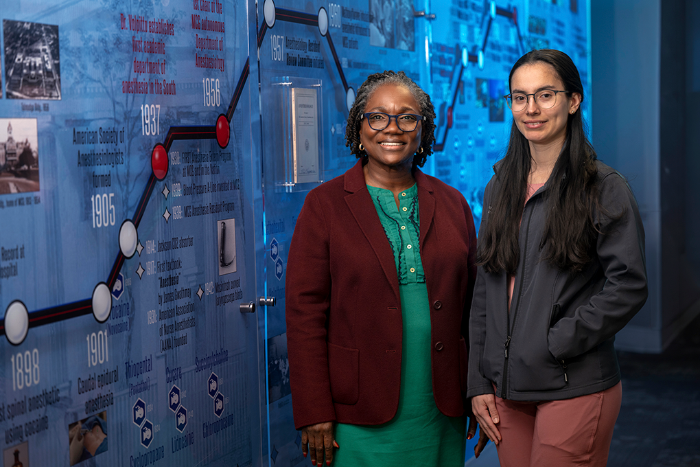 Two female anesthesiologists pose in front of a background with the history of MCG anesthesiology on it.
