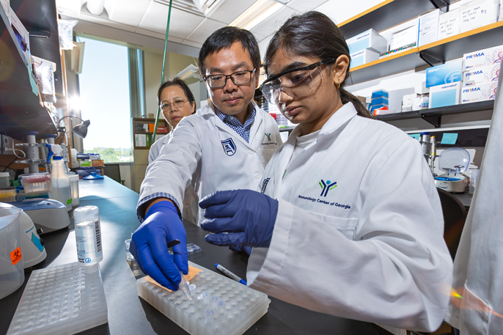 A group of four scientists gather around a lab bench to work on an experiment. Two of the scientists are working with tools, while the other two are watching.