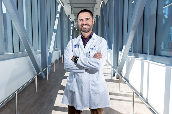A male scientist in a laboratory coat stands in a long, modern bridge connecting two scientific labs.