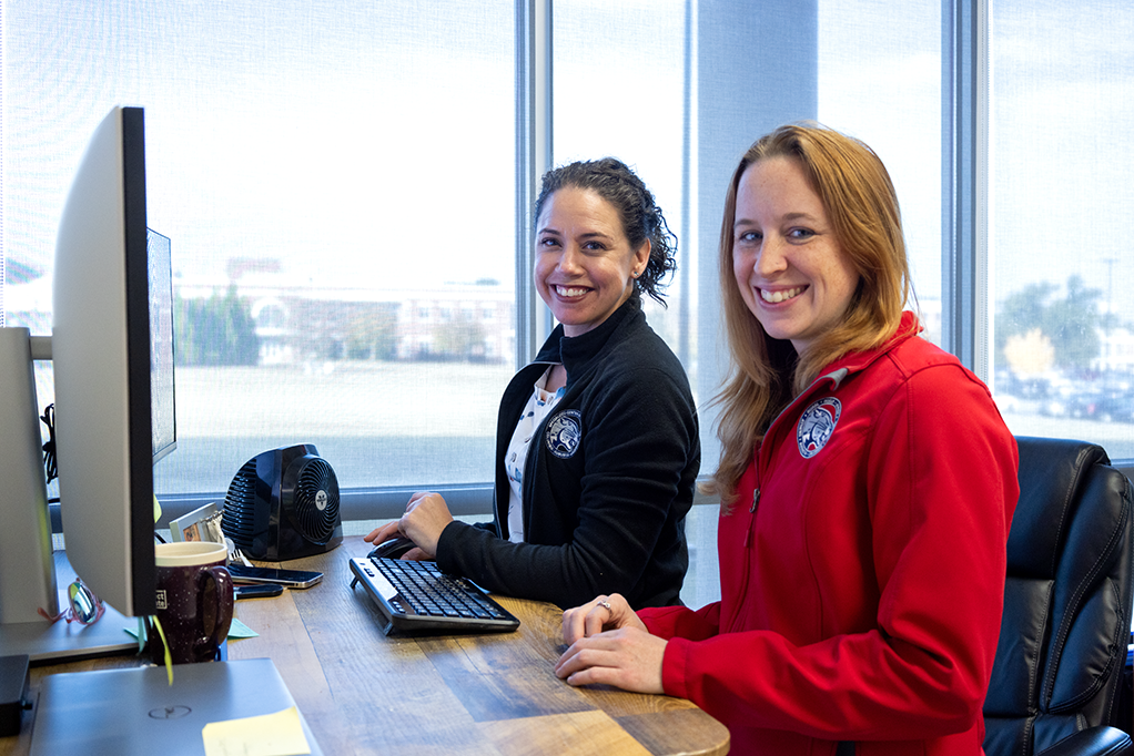 Two women smile, sitting in front of a window and computer monitor.