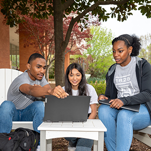 Three students sitting in front of a computer outside in the Quad
