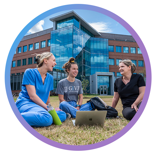 Three students are seated in the grass in front of the DCG building