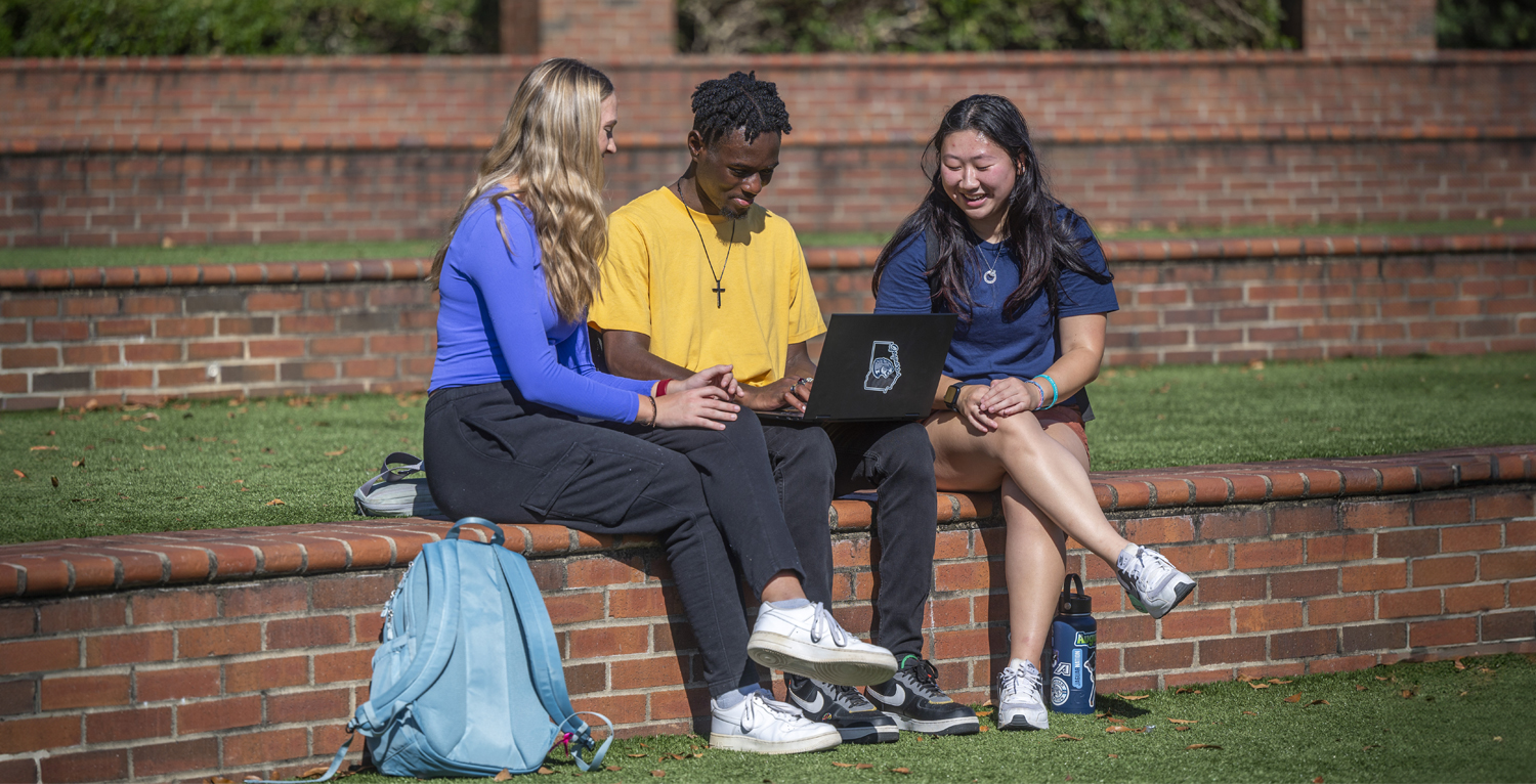 Three students sitting in the amphitheatre on the Summerville campus looking at laptop screen