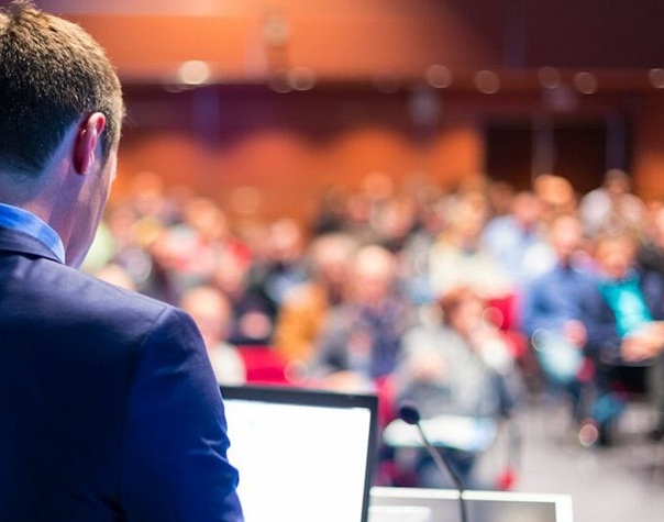 man standing speaking in front of a blurry audience