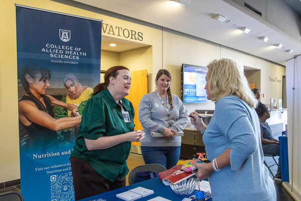 An individual speaks with two representatives at the College of Allied Health Sciences table